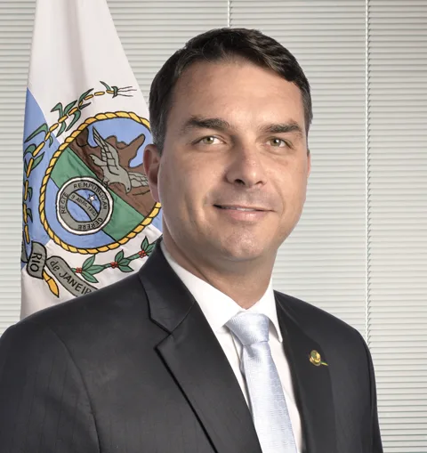 Retrato oficial de Flávio Bolsonaro, com terno escuro e a bandeira do Rio de Janeiro ao fundo.